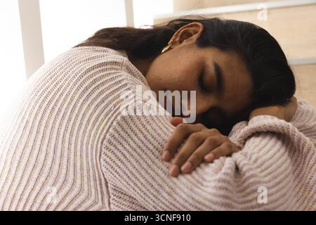 Asiatische Frau, die Strickpullover trägt, lehnt sich nach vorne mit Kopf auf Knien auf Holztreppe in der Nähe des Fensters Stockfoto
