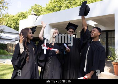 Verschiedene Klassenkameraden feiern auf dem Campus-Rasen in schwarzen Kleidern, Hüpfen und Diplomen Stockfoto