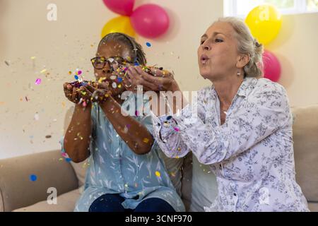 Verschiedene ältere Freundinnen blasen Papierkonfetti, während sie zu Hause mit Ballons auf der Couch sitzen Stockfoto