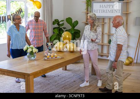 Verschiedene ältere Freunde stehen um einen hölzernen Esstisch im Wohnzimmer und bewundern Ballons Stockfoto
