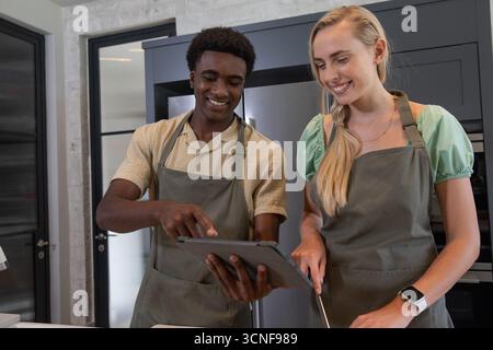 Verschiedene Freunde beugen sich an der Küchentheke über Tablette und hacken Gemüse mit einem Messer Stockfoto