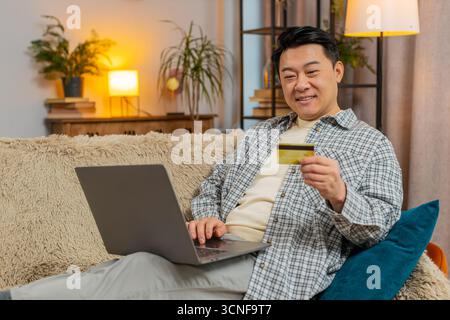 Glücklicher Mann mittleren Alters mit Kreditkarte und Laptop beim Überweisen von Geld, kauft Online-Shopping bargeldlose Bestellung Lebensmittel Lieferung zu Hause Wohnung drinnen. Der Chinese sitzt auf der Couch. Stockfoto