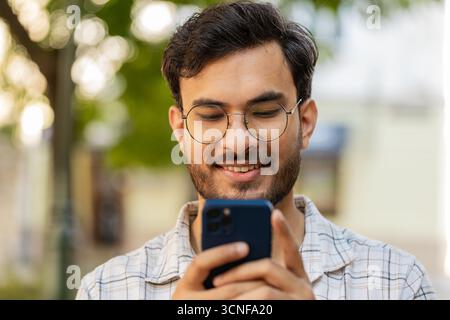 Indischer junger Mann, der per Smartphone Textnachrichten in sozialen Medien online schreibt, im Internet surft, sich entspannt, eine Pause im Freien macht. Hispanischer Kerl, der auf der Stadtstraße steht. Horizontal Stockfoto