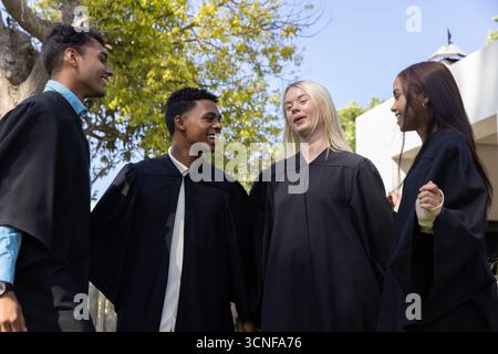 Verschiedene Klassenkameraden in schwarzen Abschlusskleidern lächeln im Innenhof, um den Abschluss zu feiern Stockfoto