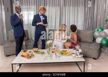 Verschiedene Hochzeitsfreunde probieren Buffet auf einem niedrigen Couchtisch in der Heim-Lounge, Cupcake-Stand Stockfoto