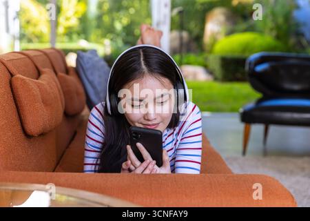 Asiatische Frau, die zu Hause auf dem orangefarbenen Sofa liegt und ein Smartphone hält und schwarze Kopfhörer trägt Stockfoto
