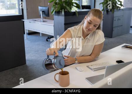 Weibliche Büroangestellte, die Kaffee aus der französischen Glaspresse in eine braune Tasse am Schreibtisch gießt Stockfoto