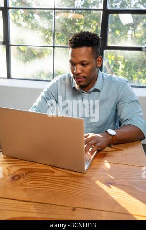 afroamerikaner, der auf silbernem Laptop am Bürotisch am Gitterfenster unter Sonnenlicht tippt Stockfoto