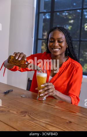 afroamerikanerin sitzt am Tisch am Gitterfenster und gießt Bier aus der Flasche Stockfoto