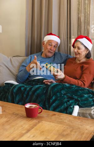 Seniorenpaar mit Santa-Hüten, die ein Geschenk auf dem Sofa im Wohnzimmer mit einem schäumenden Becher teilen Stockfoto
