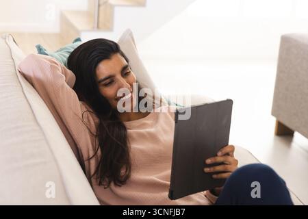 Asiatische Frau, die sich im hellen Wohnzimmer auf dem Sofa liegend, trug einen rosafarbenen Pullover und hielt ein Tablett Stockfoto