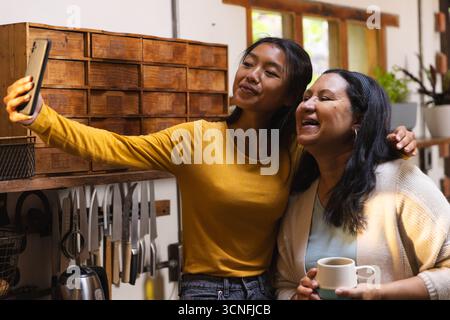 Vielfältige Mutter und Tochter machen Selfie in der Küche mit Smartphone-Becher und Messerregal Stockfoto