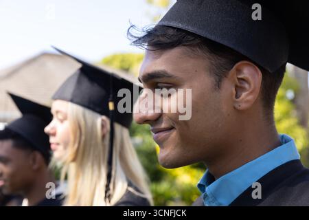 Verschiedene Absolventen stehen auf dem Campus in Schlange und tragen Mortarboards, Kleider gegen Laub Stockfoto
