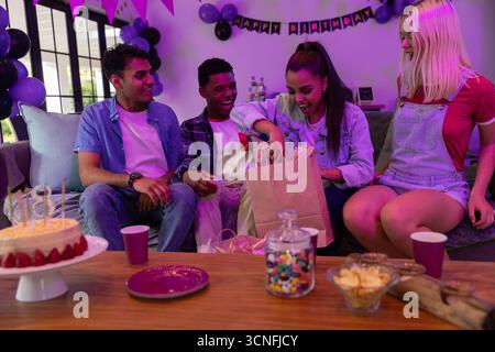 Teenager afroamerikanische Frau, die Geschenktüte auf dem Sofa öffnet, neben Geburtstagskuchen, Ballons Stockfoto