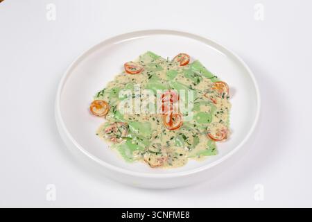 Frische grüne Ravioli aus mit Kräutern angereicherten Nudeln, serviert mit cremiger Käsesauce, halbierten Kirschtomaten und gehackten Kräutern auf einem weißen Teller. Minimal Stockfoto