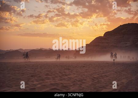 Alula, Saudi-Arabien - 04. März 2023: Reiter auf Wüstenlandschaft, mit Bergen im Hintergrund, aufgehender Sonne und orangen Wolken darüber - Beginn von en Stockfoto