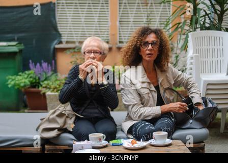 Zwei Frauen genießen an einem sonnigen Nachmittag im Freien eine Kaffeepause, umgeben von üppigem Grün und Pflanzen in einem Innenhof Stockfoto