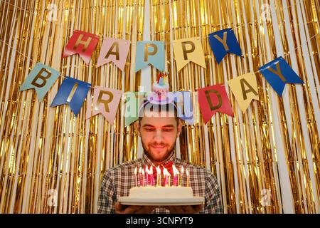 Ein Mann hält Geburtstagskuchen mit beleuchteten Kerzen, goldener Hintergrund und Happy Birthday Banner dahinter. Stockfoto