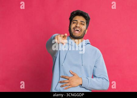 Glücklicher junger indischer Mann, der laut lacht, nachdem er lächerliche Anekdoten gehört hat, Reaktion auf lustigen Witz, unbeschwert amüsiert. Positiver Lebensstil. Arabier isoliert auf rotem Hintergrund Stockfoto