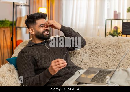 Ein indischer Mann zu Hause schrubbt die Augen mit müdem Ausdruck, während er ein Notebook benutzt, das durch eine lange Bildschirmbelichtung ermüdet ist. Ein Araber auf dem Sofa zeigt Unwohlsein und Augenanstrengung und kämpft mit digitalem Überlastungsstress Stockfoto