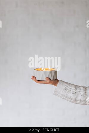 Eine Hand hält einen stilvollen, geometrischen Kerzenhalter mit zwei beleuchteten Kerzen. Der Hintergrund ist eine weiche, neutrale Farbe, die eine gemütliche Atmosphäre schafft. Stockfoto