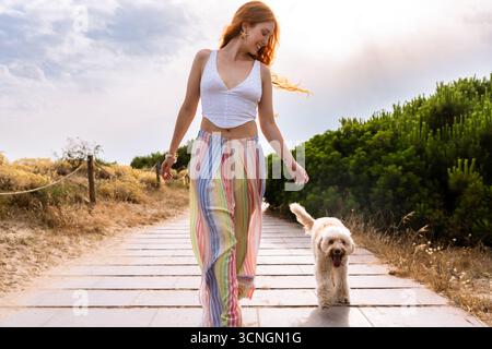 Eine rothaarige Frau und ihr goldenes Doodle spazieren auf einem sonnendurchfluteten Holzsteg durch trockenes Gras und grüne Bäume, lächeln und genießen einen unbeschwerten Sommerspaziergang Stockfoto