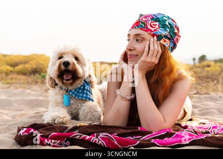 Die junge rothaarige Frau mit einem bunten Bandana liegt in warmem goldenem Licht auf einer Stranddecke, lächelt und entspannt, während sie ihr fröhliches, flauschiges Doodle kuschelt Stockfoto