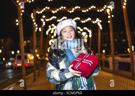 Lächelnde junge Frau, die weihnachtsgeschenk hält und die Winterferien unter dekorativen Lichtern in der Stadtstraße bei Nacht genießt Stockfoto
