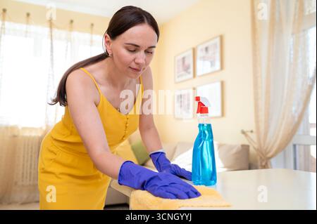 Junge freudige Frau in Handschuhen, die einen Tisch mit einem Tuch abwischt und Sauberkeit und häusliche Pflege mit einem Lächeln annimmt Stockfoto