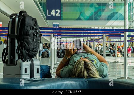 13JUN2024, Paris, Frankreich - müde weibliche Passagierin, die auf einer Couch liegt und ihr Handy mit Gepäck an der Seite im Flughafenterminal ansieht Stockfoto