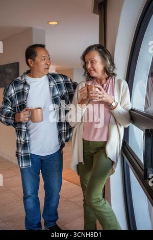 Ein älteres Paar steht zusammen im Flur neben dem Fenster und hält Kaffeetassen Stockfoto