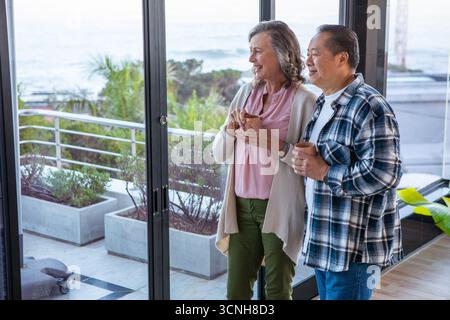 Verschiedene Seniorenpaare stehen an Schiebetüren auf dem Balkon und halten Tassen und beobachten das Meer Stockfoto