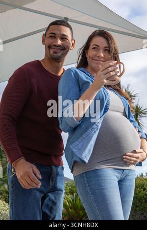 Diverse Paare mit schwangeren Partnern, die unter einem Sonnenschirm im Hinterhof stehen und Keramikbecher halten Stockfoto
