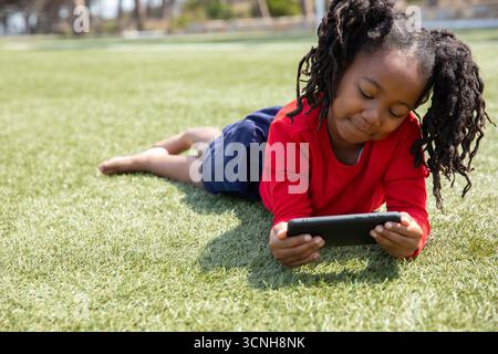 afroamerikanisches Mädchen, das mit Tablet auf Gras liegt und auf den Bildschirm im Park blickt Stockfoto