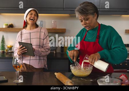 Ältere afroamerikanische Mutter mischt Teig in Schüssel als Tochter, die Tablette in der Küche hält Stockfoto