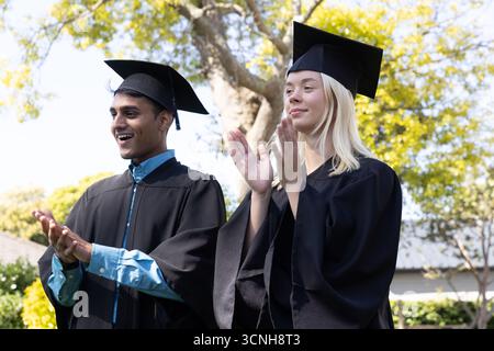 Verschiedene Absolventen in schwarzen Kleidern applaudieren den Beginn auf dem Campus-Rasen, Mörtelmützen Stockfoto