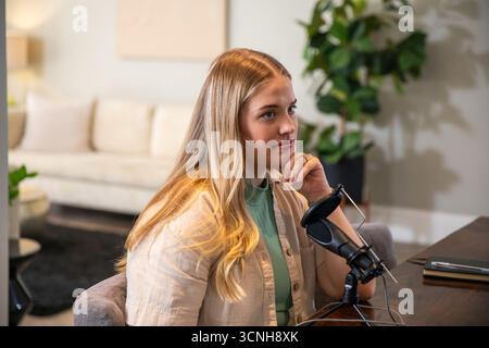 Frau, die am Holztisch sitzt und Podcast im Wohnzimmer mit Mikrofon und Popfilter aufnimmt Stockfoto