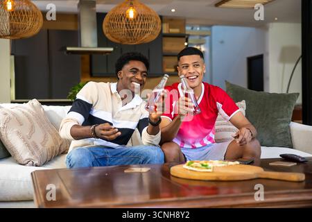 Verschiedene männliche Freunde, die Limonadenflaschen auf hellgrauem Sofa im Wohnzimmer mit Pizzabett toasten Stockfoto