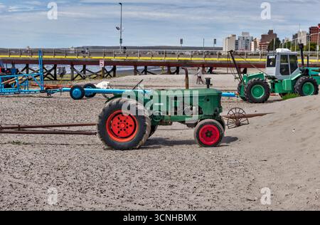 Ein grüner Traktor mit roten Rädern steht am Kieselstrand in der Nähe des Piers, der zum Starten und Bereinigen von Booten genutzt wird, mit der Skyline der Stadt Puerto Stockfoto