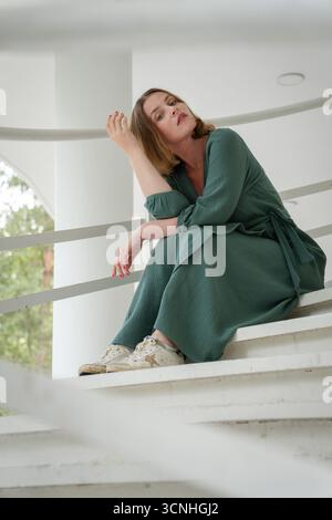 Eine Frau in grünem Kleid sitzt auf der weißen Treppe in Peredelkino und lehnt sich mit einer nachdenklichen, stilvollen Pose auf das Geländer Stockfoto