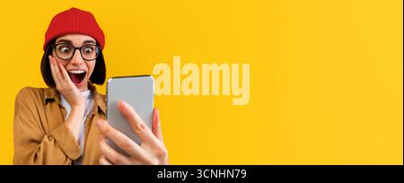 Omm. Überrascht Hipster Frau Blick auf Smartphone-Bildschirm, emotional reagiert auf gute Nachrichten, gelber Hintergrund Stockfoto