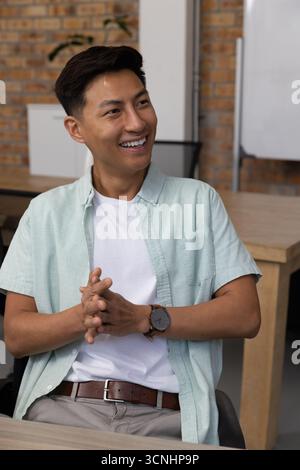 Asiatischer Mann, der lässig lächelnd und mit Händen an einem Schreibtisch im Büro sitzt Stockfoto