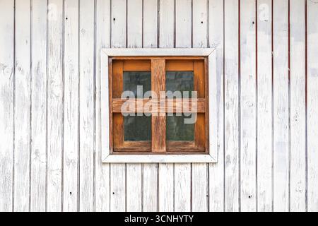 Quadratisches kleines braunes Holzfenster an der weißen Holzwand eines rustikalen Bauernhauses in der slowenischen Landschaft, einfaches alpines Architekturdetail Stockfoto
