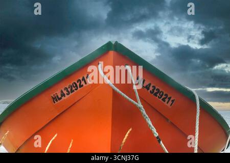 Ein dramatisches Bild eines orangen Bootes mit grüner Verkleidung sitzt ungenutzt am Sandstrand. Dunkle Wolken erheben sich über dem Himmel, während die Dämmerung den Himmel färbt und einen Drall erzeugt Stockfoto