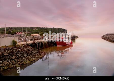 Ein leuchtend rotes Fischerboot liegt bei Flut in Alma, New Brunswick, Kanada, bei Sonnenaufgang sicher an. Der pastellfarbene Himmel schafft eine friedliche Atmosphäre Stockfoto