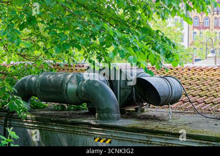 Auf einem Betondach ist ein großes Metallgehäuse für Heizungs- und Klimatechnik mit einem Auspuffrohr mit Gitterkappe verbunden. Mehrere Gebäudekanäle versorgen die Einheit. Sauberes industrielles Gerät Stockfoto