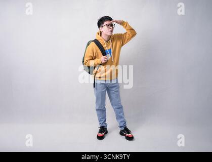 Junger Mann mit Rucksack in gelbem Hoodie, der Pose im Studio-Porträt grüßt Stockfoto