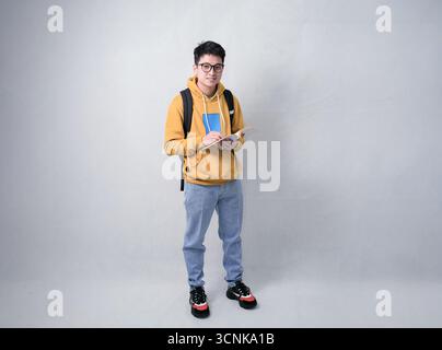 Ein junger Schüler steht in einem Studio, trägt einen Senfpullover und einen Rucksack, während er liest und Notizen macht. Lässig Stockfoto