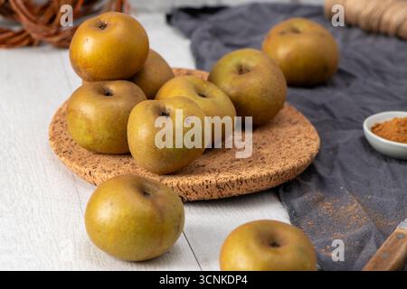 Eine Stillleben-Komposition aus rustikalen grünen und gelben Äpfeln, von denen einige auf einer Korkmatte gestapelt und andere auf einem weißen Holztisch verstreut sind. Eine kleine Schüssel Stockfoto