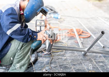 Ein Bauarbeiter in Schutzkleidung und blauem Schweißhelm schneidet auf einer Baustelle ein Edelstahlrohr mit einem Winkelschleifer. Stockfoto